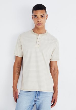 Chemise henley à manches courtes, rayée crème, avec trois boutons et un col côtelé. Portée avec un jean en denim bleu clair.