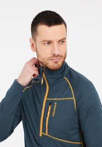 Tamsiai mėlynas megztinis su aukštu apykakle, turintis oranžinį siuvinėjimą ir užtrauktuką krūtinės kišenėje. Pagamintas iš tekstūruotos medžiagos, skirtas sluoksniavimui.