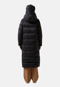 Manteau noir long en duvet avec capuche, arborant des sections matelassées horizontales et une texture lisse et brillante, associé à un pantalon marron décontracté.