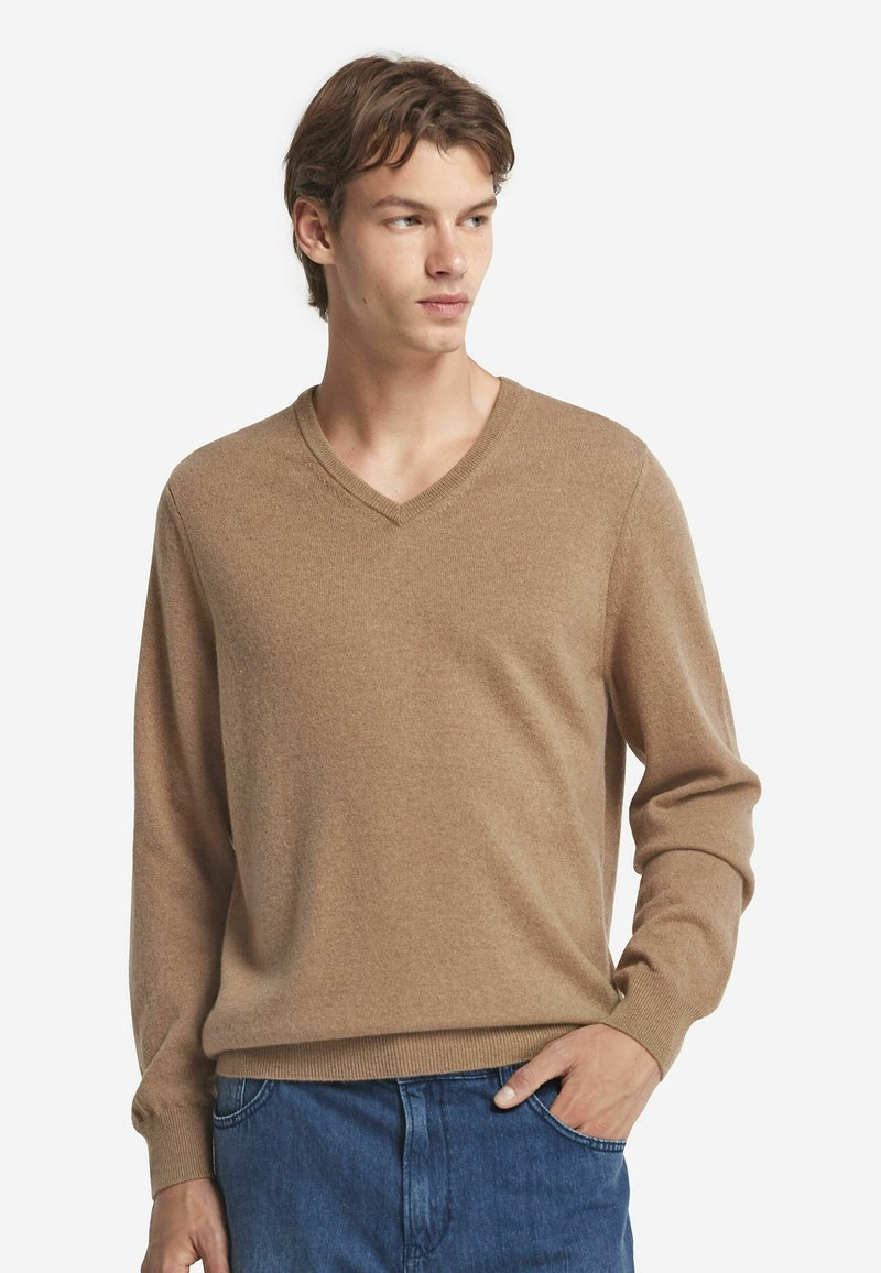 Brązowy sweter w serek, wykonany z miękkiego materiału, z długimi rękawami, ściągaczami na mankietach i u dołu. Noszony na niebieskich dżinsach, model pozuje z jedną ręką w kieszeni.