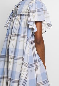 Osoba v volných, světle modrých a bílých kostkovaných šatech s krátkými, nabíranými rukávy a nařasenými detaily.
