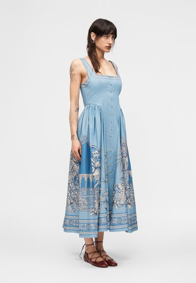 Γυναίκα με τατουάζ που φοράει μίντι φόρεμα μπλε αμάνικο με λευκά λουλουδάτα σχέδια και κουμπιά, συνδυασμένο με καφέ δερμάτινα παπούτσια με κορδόνια.