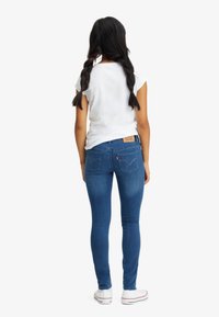 Jeans ajustados de color azul hechos de mezclilla, con una etiqueta en la cinturilla y un diseño clásico de bolsillo trasero, combinados con una camiseta blanca de manga corta.