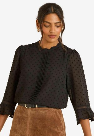 Schwarze Bluse mit strukturierten Punkten, Puffärmeln und einem geschwungenen Ausschnitt, ausgestattet mit vorderen Falten und durchscheinenden Stoffakzenten.