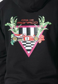 Felpa nera con cappuccio caratterizzata da una grafica di una macchina da corsa rossa con il numero "1" su di essa, incorniciata da foglie e fiori verdi, su uno sfondo a scacchi e un triangolo rosa.