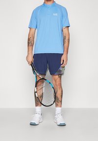 Man i blå EA7 sporttröja och marinblå shorts som håller en svart och blå tennisracket, med synliga tatueringar på armar och ben.