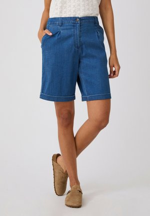 Personne portant un short en jean bleu avec ourlets, un haut sans manches de couleur claire et des chaussures beiges à enfiler, debout avec une jambe croisée sur l'autre.