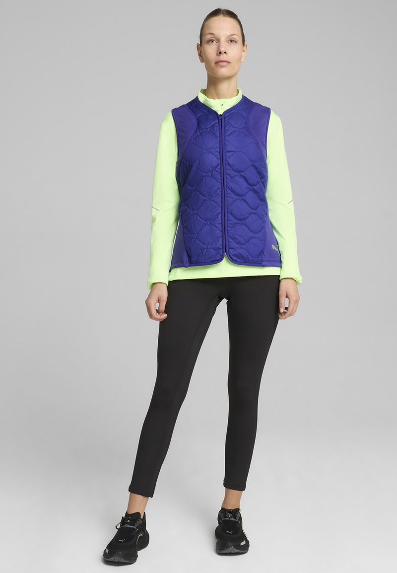 Violet komplikuotas liemenė su aukšta apykakle, dėvima ant ry�škiai žalia ilgomis rankovėmis marškinėlių. Sujungta su juodomis tampančiomis kelnėmis ir sportiniais batais.