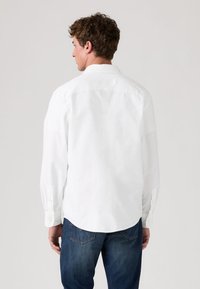 Cămașă albă cu mânecă lungă și guler cu nasturi, având o textură fină de bumbac. Asortată cu blugi din denim albastru închis, prezentând o vedere din spate.