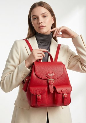 Jonge vrouw in beige blazer en zwarte coltrui, met een rode leren rugzak met gespsluitingen en voorvakken.
