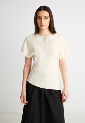 Fiatal nő hosszú, sötét hajjal, texturált törtfehér rövid ujjú felsőt és magasderekú fekete szoknyát viselve, egyszerű háttér előtt áll.