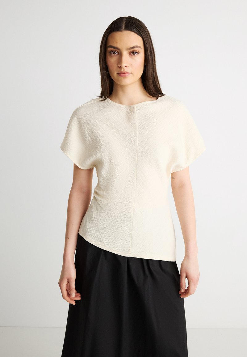 Νεαρή γυναίκα με μακριά σκούρα μαλλιά, φορώντας υφασμάτινη κοντομάνικη μπλούζα εκρού και μαύρη φούστα με ψηλή μέση, στέκεται μπροστά σε ένα απλό φόντο.