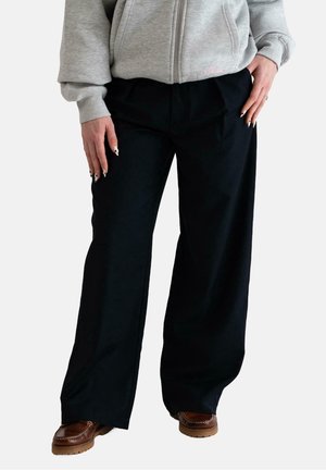 Persoon gekleed in zwarte wijd uitlopende broek, bruine schoenen en een grijze zip-up hoodie met handen rustend nabij de heupen.