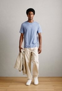 Tricou scurt de culoare albastru deschis, pantaloni crem și adidași albi. Modelul ține o jachetă bej, stând în fața unui fundal neutru.