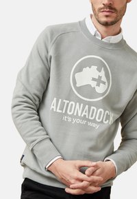 Sudadera gris hecha de un tejido suave que presenta un logotipo circular con la silueta de un mapa y el texto "ALTONADOCK es tu camino" en blanco.