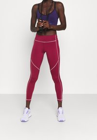 Leggings bordeaux a vita alta, aderenti, con cuciture laterali argento in contrasto, abbinati a un reggiseno sportivo viola e scarpe da ginnastica bianche.