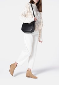 Botas de ante marrón claro hasta el tobillo con un tacón bajo, combinadas con pantalones blancos ajustados y un suéter de punto de cable color crema con detalles de flecos. Bolso negro de hombro.