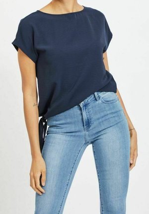 Femme portant un haut bleu marine à manches courtes noué sur le côté et un jean skinny taille haute bleu clair, debout les mains détendues.