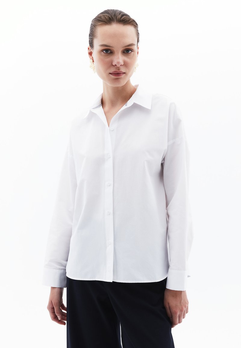 Witte blouse met een klassieke kraag, lange mouwen en een licht gebogen zoom. De stof lijkt glad met een frisse textuur.