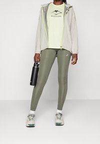 Persona vistiendo una camiseta ASICS Fujitrail verde claro, una chaqueta ligera, leggings verde olivo, zapatillas, sosteniendo una botella de agua negra, de pie sobre un fondo blanco.