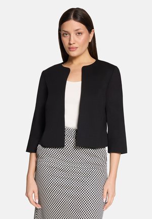 Vrouw met een zwarte open jas, wit topje en een zwart-witte rok met patroon, staand tegen een effen lichte achtergrond.