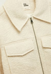 Fleece-foret cremet jakke med lynlås, spids krave og to forlommer. Har et blødt struktureret stof og syet detaljer.