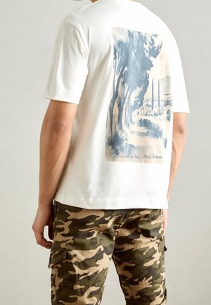Person trägt ein weißes T-Shirt mit blauem Aquarell-Landschaftsprint und beige-grünen Camouflage-Cargohosen, Sicht von hinten.
