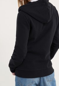 Sudadera negra con capucha, puños y dobladillo acanalados, textura suave y ajuste casual. Combinada con jeans de mezclilla azul claro.