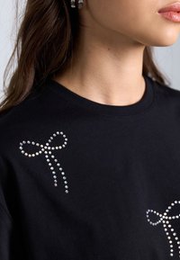 Musta puuvillapaita, jossa on kaksi kiiltävästä strassista tehtyä nauhasuunnitelmaa edessä, sileä pinta, pyöreä kaula-aukko.