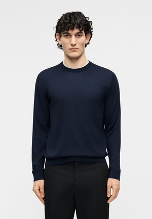 Fiatal férfi göndör sötét hajjal, sötétkék kereknyakú pulóvert és fekete nadrágot viselve, egyszínű fehér háttér előtt áll.