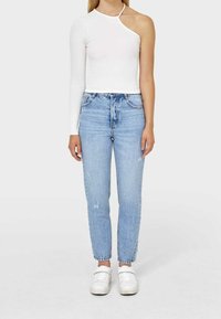 White asymmetrical top with one long sleeve, paired with light blue high-waisted jeans and white sneakers, featuring distressed accents.