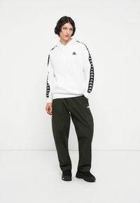 Sudadera blanca con logo negro y rayas laterales, combinada con pantalones cargo verdes oliva. El modelo lleva zapatillas negras. Textura de tela suave.
