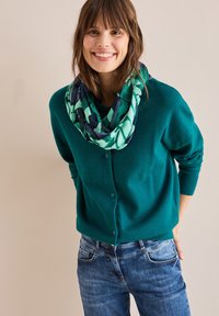 Tyrkysový pletený kardigan s knoflíkovým zapínáním, nošený přes vzorovaný zelený a námořnický šál ve tvaru infinity, spárovaný s modrými džíny.