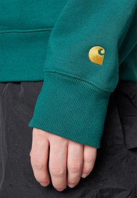 Sudadera verde con puños de canalé y un parche de logo dorado. Primer plano de la manga y la mano, mostrando la textura del material y los detalles de la costura.