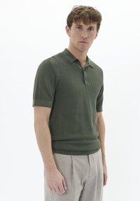 Polo cu mânecă scurtă, de culoare verde închis, cu un model texturat în zigzag, având guler și manșete ribbate, asortat cu pantaloni ușori, pliați.