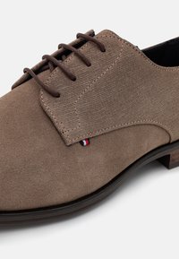 Chaussure habillée à lacets pour homme en daim marron avec panneaux latéraux texturés, lacets marron foncé et petite étiquette en tissu rouge, blanc et bleu près de la couture.