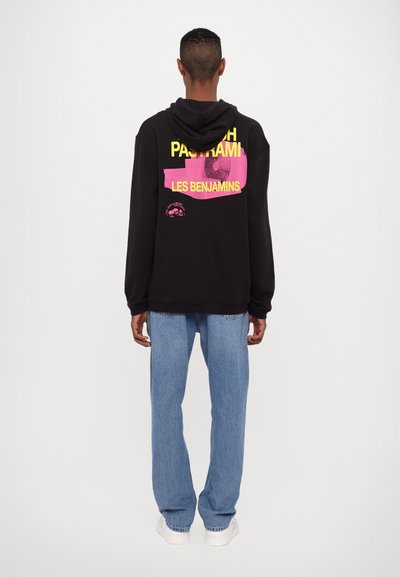 Black hoodie with a front pocket, featuring bright yellow and pink graphic text on the back. Paired with light blue jeans and white sneakers.