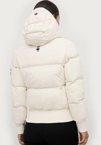 Giacca imbottita di colore bianco sporco con colletto alto e grande cappuccio imbottito. Presenta polsini e orlo a coste, con un logo nero accentato sulla schiena.