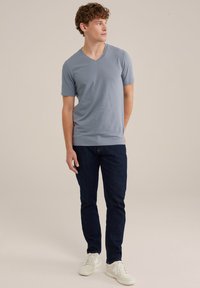 Svetlo modra v-vratna majica iz mehke tkanine s kratkimi rokavi, oblečena z temnimi kavbojkami in belimi supergami, stoji na nevtralni podlagi.