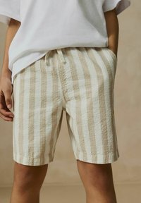 Shorts rayés beige clair et blancs en tissu texturé. Dotés d'une taille élastique avec cordon de serrage et de poches latérales.