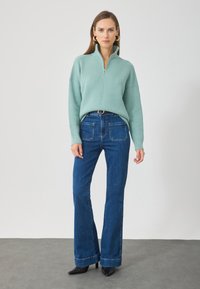 Miętowozielony sweter w prążki z ćwierć zamkiem, zestawiony z niebieskimi spodniami dzwonami z dwoma tylnymi kieszeniami i paskiem w talii. Czarne obcasy.