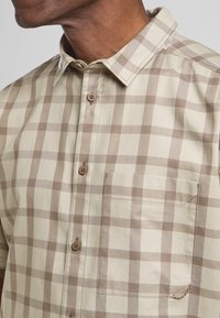 Camicia a maniche corte, a scacchi beige e marrone, con colletto a punta, bottoni in legno e tasca sul petto con cuciture decorative.