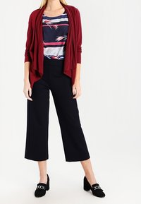 Burgundská cardigan s otevřeným předním dílem přes námořnický vzorovaný top, doplněná černými širokými kalhotami a černými mokasínami s řetízkovými detaily.
