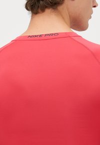 Rde majica kratkih rokavov Nike Pro, s prijetno teksturo in prilegajočim dizajnom. Črn logotip "NIKE PRO" je postavljen na zadnjem izrezu ovratnika.