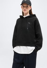 Une jeune personne porte un sweat à capuche noir avec un logo de crocodile, une chemise blanche en dessous, un pantalon foncé et une casquette noire devant un fond clair uni.