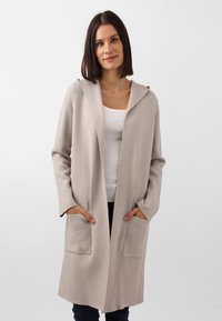 Cardigan beige con cappuccio, dalla texture morbida, apertura frontale e due tasche anteriori. Maniche lunghe e vestibilità rilassata. Abbinato a un top bianco.