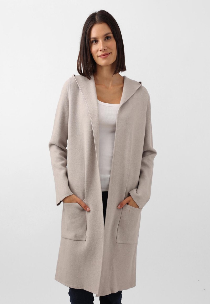 Cardigan beige con cappuccio, dalla texture morbida, apertura frontale e due tasche anteriori. Maniche lunghe e vestibilità rilassata. Abbinato a un top bianco.
