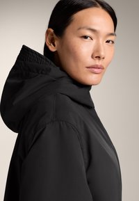 Veste noire à capuche avec une finition lisse et un col matelassé. Gros plan sur le profil, mettant en avant la texture du tissu et les lignes nettes.