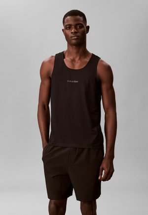 Mężczyzna stojący z rękami w kieszeniach, ubrany w czarny top bez rękawów marki Calvin Klein i czarne szorty, na tle jednolitego szarego tła.