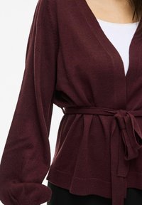 Cardigan lavorato a maglia bordeaux con scollatura a V profonda, vita con cintura e maniche a palloncino. Motivo testurizzato lungo le maniche e orlo a coste.